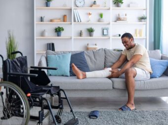Millennial black man having plastered broken leg, suffering from pain on sofa at home, wheelchair standing nearby