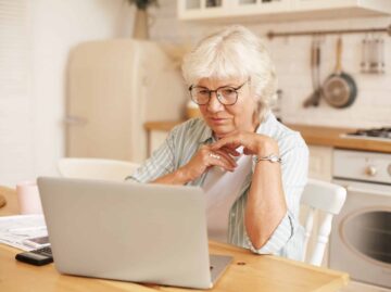 Modern technology, old people and retirement. Gray haired female pensioner in glasses filling in loan application form online, sitting in front of laptop, reading information with serious focused look
